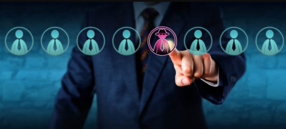 A person in a suit points to an icon of a figure wearing a hat and mask among several generic human icons, symbolizing effective threat detection and identification of a suspicious or malicious individual.