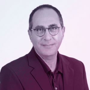 A middle-aged man with short dark hair, glasses, and a slight smile, wearing a dark blazer over a collared shirt, poses against a plain light background—ideal for discussing cyber crime on the Cyber Crime Junkies Podcast.