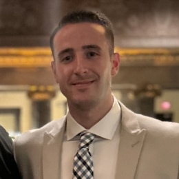 A man in a light-colored suit and checkered tie stands indoors, smiling slightly, with a blurred ornate background behind him at FutureCon OC 2024.