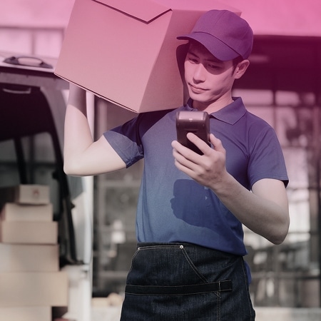 A delivery worker in a blue uniform, displaying true hospitality, holds a package on his shoulder and checks a handheld device, standing next to a vehicle with more boxes.